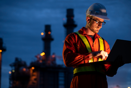 Man wearing hard hat looking at laptop standing in front of power station at night